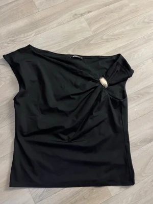 Svart asymmetrisk topp med brosch - Snygg svart topp med asymmetrisk halsringning och dekorativ broschdetalj vid axeln. Materialet känns mjukt och följsamt. Från Gina tricot. Katt finns i hemmet!