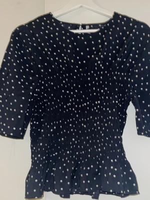 Svart prickig blus med trekvartsärm - Snygg svart blus med vita prickar och trekvartsärm. Blusen har rund halsringning och smockad midja som ger en feminin siluett. Tillverkad i ett lätt och luftigt material som känns skönt mot huden. Perfekt för dig som gillar trendiga mönster. Knappt använt och är inifrån JYOTHI storlek L men passar mig som är S men funkar bättre som M 