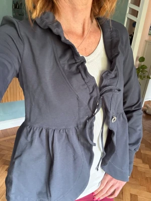 Mörkgrå kofta med volang och knyt - Mörkgrå kofta med volangkant längs öppningen och knytband framtill. Koftan har långa ärmar och en lätt rynkad midja som ger en feminin siluett. Materialet ärbommulls  trikå. Mycket  mjukt och bekvämt, perfekt för att lägga över en topp. Storlek small. Obs knappen har lossnat men lätt att sy fast. Jag saknar dock tråd i rätt nyans. Men knappen hänger med i en liten säkerhetsnål 