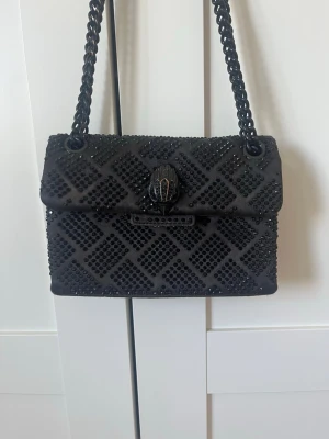 Svart axelväska från Kurt Geiger - Säljer en elegant svart axelväska från Kurt Geiger med kedjerem och dekorativt mönster av glittriga svarta stenar. Väskan har ett unikt spänne med ormhuvud och ett rutigt mönster på hela ytan. Perfekt för dig som vill sticka ut med en lyxig accessoar.