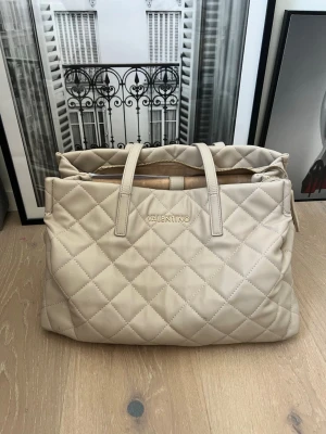 Beige quiltad handväska från Valentino - Stilren beige handväska från Valentino med quiltat mönster och dubbla handtag. Stor och perfekt för skolan. Väskan har guldfärgad logga framtill och dragkedja upptill. Tillverkad i mjukt konstläder med lyxig känsla och rymlig insida för alla dina essentials.