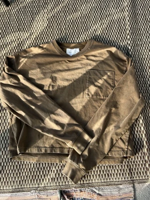 Beige croppad långärmad topp H&M Studio - Säljer en beige croppad långärmad topp från H&M Studio S/S 2025. Toppen har en enkel design med rund halsringning och en bröstficka. Perfekt för dig som gillar stilrena plagg med lite edge.
