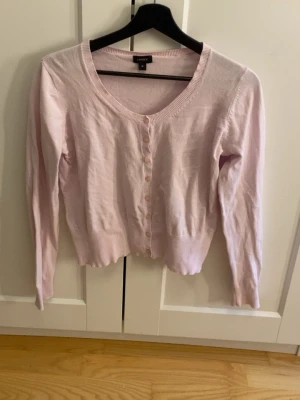 Ljusrosa kofta från Lindex - Söt ljusrosa kofta från Lindex med rund halsringning och knappar framtill. Långärmad modell i tunt, mjukt material med ribbade muddar vid ärmslut och nederkant. Perfekt att slänga över en topp för en enkel och stilren look.