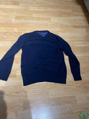 Mörkblå stickad tröja Tommy Hilfiger - Snygg mörkblå stickad tröja från Tommy Hilfiger med diskret logga broderad på ärmen. Tröjan har långa ärmar och ribbade muddar vid ärmslut och nederkant. Perfekt till jeans eller chinos för en clean look. Sitter som en s/m beroende på önskad passform. 