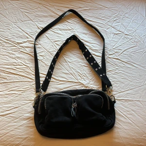 Svart axelväska - Cool svart axelväska i mockaimitation från Gina Tricot med två stora framfickor och silvriga dragkedjor. Väskan har en bred axelrem med nitar och karbinhakar i metall. Perfekt för att få plats med allt du behöver 🖤 OBS! Det långa bandet ingår ej!