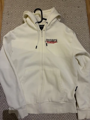 Vit zip hoodie från Hoodrich - Säljer en vit hoodie med dragkedja från Hoodrich. Hoodien har huva med dragsko, fickor fram och tryck med Hoodrich-logga både på bröstet och ryggen. Det finns även tryck på ena ärmen. Materialet är mjukt och skönt, perfekt för en avslappnad streetwear-look.