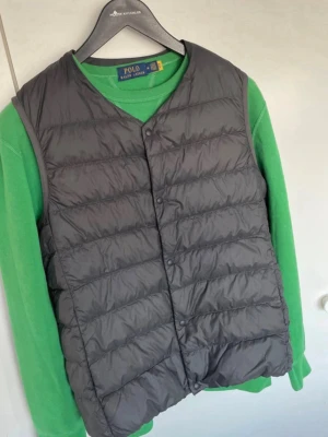 Grå dunväst från Uniqlo’s  - Snygg grå dunväst från Uniqlo med knäppning framtill och klassisk quiltad design. Västen har V-ringning och är lätt vadderad, perfekt att slänga över en hoodie eller tröja för extra värme. Enkel och stilren look som funkar till det mesta.