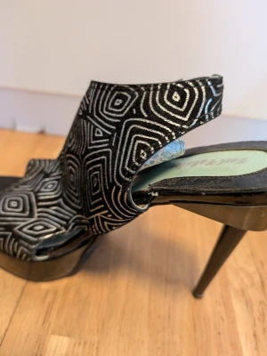 Svarta platåsandaletter med mönster - Unika svarta platåsandaletter från Feel Fabulous med höga klackar och grafiskt silvermönster. Skorna har öppen tå och en cool, modern design. Perfekta för dig som vill sticka ut med din stil.Använda vid några få tillfällen.Klackarna är ca 9-10 cm.Börjar "flagna" inuti, se bild.