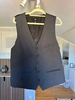 Mörkgrå kostymväst från H&M - Stilren mörkgrå kostymväst från H&M med klassisk passform. Västen har tre fickor framtill, knäppning med fyra knappar och är helfodrad i svart tyg. Perfekt att matcha med kostymbyxor för en dressad look.