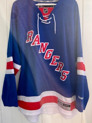New York Rangers - NHL - Officiell hockeytröja - Säljer en New York Rangers-matchtröja. Tröjan är i väldigt bra skick utan några defekter som jag kan se. Officiell NHL-produkt från Reebok. Inget namn eller nummer på ryggen.  Storlek: Large  #SHL #Sport #Hockey #Lundqvist #Sverige #Hockeybilder