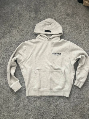 Grå Essentials Fear of God hoodie - Snygg ljusgrå hoodie från Fear of God Essentials med svart logga på bröstet och stor text på ryggen. Klassisk känguruficka framtill och huva. Perfekt för en clean och avslappnad streetwear-look.