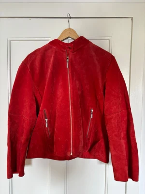 Röd skinnjacka med dragkedja - Cool röd skinnjacka i mocka-look med rak passform och ståkrage. Jackan har två snedställda fickor med dragkedjor framtill och en snygg, minimalistisk design utan synliga loggor. Perfekt för dig som vill sticka ut med färg.