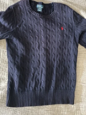 Mörkblå kabelstickad tröja Polo Ralph Lauren - Kabelstickad mörkblå tröja från Polo Ralph Lauren med rund halsringning och klassisk röd broderad logga på bröstet. Tröjan har långa ärmar och breda ribbade muddar vid ärmslut och nederkant. Perfekt för en stilren och tidlös look.