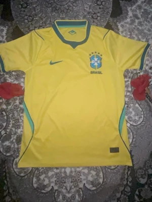 Brasilien Nike fotbollströja gul - Gul Brasilien fotbollströja från Nike med gröna och blå detaljer, CBF-emblem och Nike-logga på bröstet. Klassisk rund halsringning och korta ärmar. Perfekt för dig som älskar fotboll och vill visa ditt stöd för Brasilien.