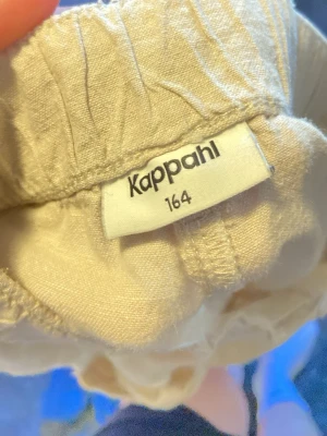 Beiga linnebyxor från Kappahl - Snygga och luftiga beiga linnebyxor från Kappahl i storlek 164. Byxorna har resår i midjan och är raka i modellen, perfekta för en avslappnad och stilren look. Tillverkade i linne för en skön känsla under varma dagar.