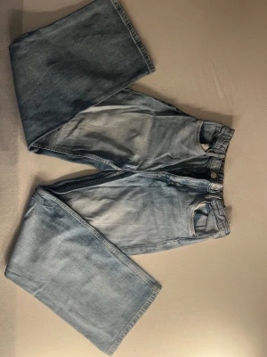 Ljusblåa vida jeans med hög midja - Säljer ett par ljusblå jeans med hög midja och vida ben. Klassisk femficksmodell med dragkedja och knapp framtill. Jeansen har en avslappnad passform och är tillverkade i denim. Perfekta för en trendig och chill look.