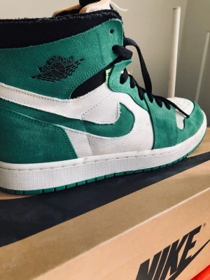Nike Air Jordan 1 Retro High grön/vit - Säljer ett par Nike Air Jordan 1 Retro High sneakers i grön och vit mocka med svarta detaljer och klassisk Air Jordan-logga på sidan. Skorna har snörning och en hög siluett som ger en ikonisk streetwear-look. Perfekta för dig som vill sticka ut med stil.
