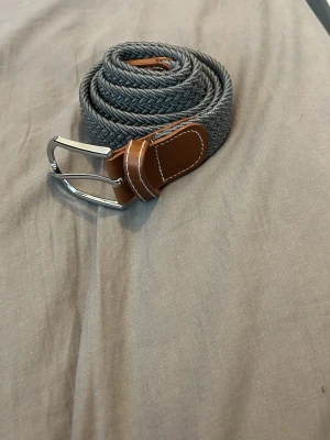 Grått flätat bälte - Snyggt grått flätat bälte med bruna skinn-detaljer och silverfärgat spänne. Perfekt accessoar för att lyfta din outfit och funkar till både jeans och chinos. Bältet har en stilren look och passar till många olika stilar.