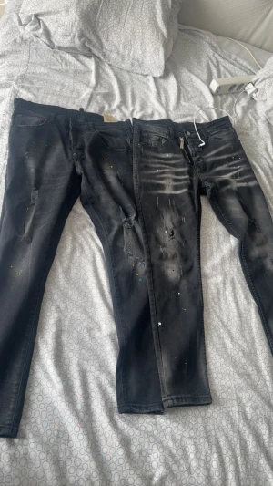 Svarta  jeans med märket dqsuared2 - Säljer ett par svarta  jeans från Dsquared2 med slitningar, färgstänk i blått och gult samt distressed detaljer på benen. Jeansen har fem fickor och en klassisk knappgylf. Perfekta för en edgy och trendig streetwear-look.