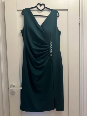 Mörkgrön omlottklänning  - Stilren mörkgrön klänning med omlott-effekt och dekorativa silverfärgade knappar i sidan. Ärmlös design med v-ringning och lätt drapering framtill. Perfekt för dig som gillar klassiska och eleganta plagg.