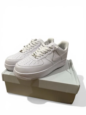 Nike Air Force 1 vita sneakers - Klassiska Nike Air Force 1 sneakers i helvitt läder med perforerad tå, vit sula och vita skosnören. Ikonisk Swoosh-logga på sidan och metallbricka på snörningen. Perfekt för en clean och stilren look. Du vet varför priset är lågt