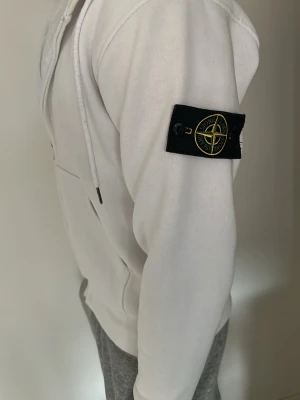 Vit zip-hoodie från Stone Island - Vit zip-hoodie från Stone Island med klassisk logotyp-patch på vänster ärm. Hoodien har dragsko i huvan, ribbade muddar och fickor framtill. Använd få gånger😇