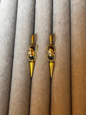 Guldpläterade örhängen med strass och spikes - Säljer ett par trendiga guldpläterade örhängen med små ringar, gnistrande strassdetaljer och hängande konformade spikes. Örhängena har en edgy vibe och passar perfekt för dig som gillar statement-accessoarer. Materialet är metall med guldfinish och glittrande stenar.