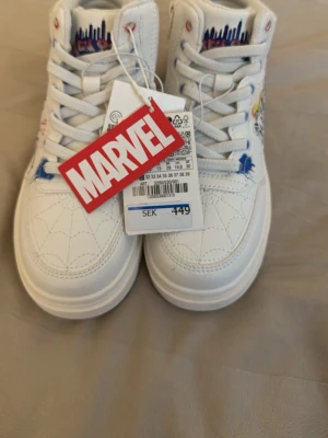 Marvel Miles Morales vita sneakers - Säljer ett par vita sneakers med Marvel Miles Morales-tema. Skorna har coola grafiska tryck med spindelnät, Miles Morales och textdetaljer på sidorna. De är tillverkade i syntetmaterial och har vita snören samt en chunky sula. Perfekta för dig som gillar superhjältar och streetstyle.