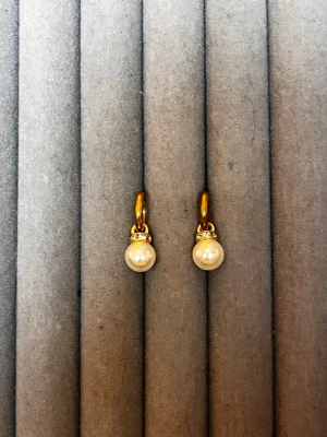 Guldpläterade örhängen med pärlor - Snygga örhängen med guldfärgade ringar och vita pärlor. Ovanför pärlan sitter en liten rad med gnistrande stenar som ger extra bling. Perfekta för dig som gillar klassisk men trendig stil. Materialet ser ut att vara metall med guldplätering och konstpärlor.