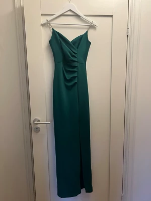 Grön långklänning med slits - Elegant grön långklänning med smala axelband och v-ringad front. Klänningen har draperad detalj framtill och en diskret slits som ger en snygg siluett. Perfekt för dig som vill sticka ut med en stilren look. Materialet känns mjukt och följsamt.