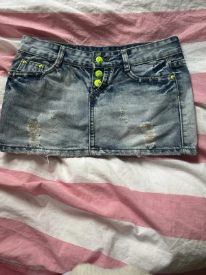 Kort jeanskjol med slitningar och neonknappar - Snygg vintage jeanskjol  i ljusblå denim med slitna detaljer framtill och bak. Kjolen har neonfärgade knappar , samt bakfickor varav en med dragkedja. Säljer då den blivit för liten❣️
