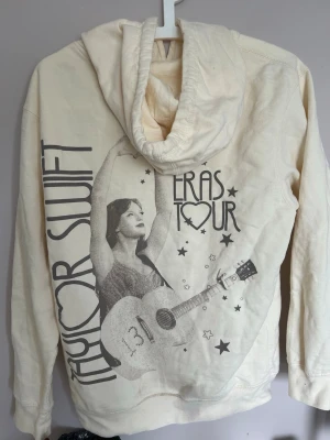 Taylor Swift Eras Tour hoodie beige - Beige hoodie med stor Taylor Swift Eras Tour-print på ryggen och mindre Taylor Swift-tryck fram. Hoodien har huva med dragsko och en stor magficka. Tillverkad i mjukt bomullsmaterial, perfekt för dig som älskar musik och vill ha en unik merch. Defekt, ena snöret är fransigt.