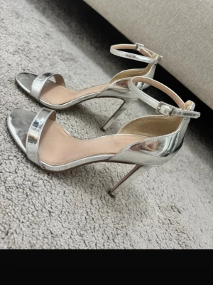 Silvriga metallicpumps med smal rem 60kr  - Snygga pumps i metallic-silver med smal rem över tårna och runt ankeln. Klackarna är smala och höga för en elegant look. Skorna har öppen tå och spänne vid ankeln. Perfekta för att lyfta din outfit med en glansig touch.