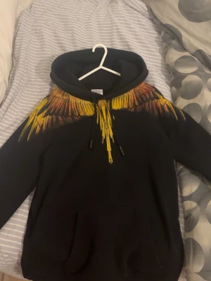 Svart hoodie Marcelo Burlon( ÄKTA ) - ( Nyskick )Snygg svart hoodie från Marcelo Burlon County of Milan med ikoniskt vingtryck i gult och orange över axlar och bröst. Hoodien har huva med dragsko och stor känguruficka framtill. Perfekt statement-plagg för dig som vill sticka ut. Passar S och m. NY PRIS 3599 kr kvitto och qr kod finns hör av mer vid fler funderingar 