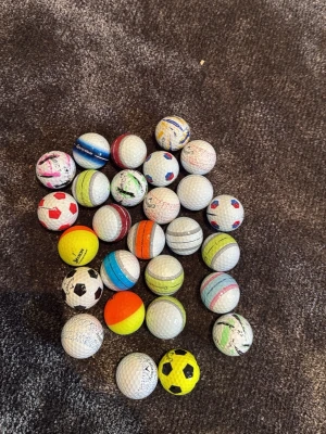 Samling med olika bollar - En samling med små bollar i olika färger och mönster, bland annat golfbollar, tennisbollar och bollar med fotbollsmönster. Färgerna varierar mellan vitt, gult, orange, blått, rött och svart. Perfekt för lekar, träning eller samling.