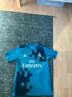 Real Madrid bortatröja Ronaldo #7 - Säljer en blå Real Madrid bortatröja från Adidas med Ronaldo och nummer 7 på ryggen. Tröjan har digitalt pixelmönster i svart, klubbmärke och Adidas-logga på bröstet samt Fly Emirates-tryck. Perfekt för fotbollsträning eller att bära som supporter.