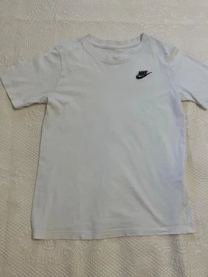 Vit t-shirt från Nike med logga - Klassisk vit t-shirt från Nike med svart broderad logga på bröstet. T-shirten har rund halsringning och korta ärmar. Tillverkad i mjuk bomull för en skön känsla och enkel stil. 12-13 år