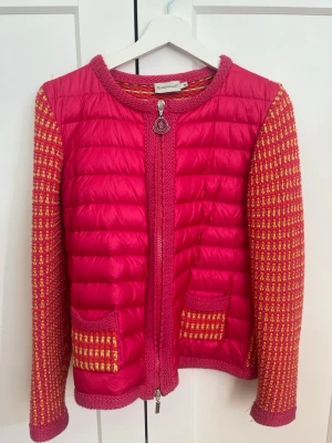 Rosa pufferjacka från Moncler - Säljer en unik rosa pufferjacka från Moncler med stickade ärmar och detaljer i rosa och gult. Storleken är M men sitter jättebra på mig som är XS/S. Jackan har två framfickor, rund halsringning och dragkedja med Moncler-logga. Materialet är en mix av quiltad nylon och stickat tyg för extra stilpoäng.