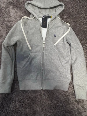 Grå hoodie från Polo Ralph Lauren - Snygg grå hoodie från Polo Ralph Lauren med dragkedja framtill, vit dragsko och klassisk broderad logga på bröstet. Mjuk bomullsblandning och huva gör den perfekt för en chill och stilren look.