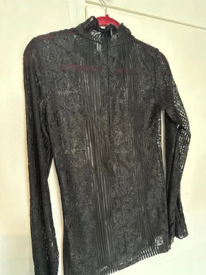 Svart spetsblus med hög krage - Snygg svart blus i transparent spets med lång ärm och hög krage. Blusen har ett detaljerat blommönster och dragkedja bak. Perfekt för dig som gillar en edgy och elegant stil. Materialet är tunt och stretchigt, vilket ger en skön passform och style 🌸
