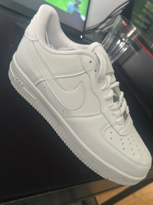 Nike Air Force 1 vita sneakers - Klassiska Nike Air Force 1 sneakers i helvitt läder med vit sula och vita skosnören. Ikonisk swoosh-logga på sidan och perforerade detaljer på tån för extra ventilation. Perfekt för dig som gillar en clean och tidlös look.