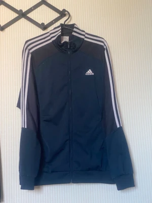 Marinblå Adidas träningsjacka - Snygg marinblå träningsjacka från Adidas med klassiska vita ränder längs ärmarna och dragkedja framtill. Jackan har hög krage, muddar vid ärmslut och nederkant samt Adidas-logga på bröstet och ryggen. Perfekt för sport eller chill.