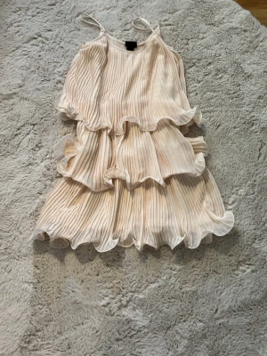 Plisserad volangklänning från H&M - Supersöt ljusbeige klänning från H&M med tunna axelband och flera lager volanger. Klänningen är plisserad och har en luftig, flowy känsla som ger en romantisk vibe. Perfekt för dig som gillar en feminin och trendig stil.