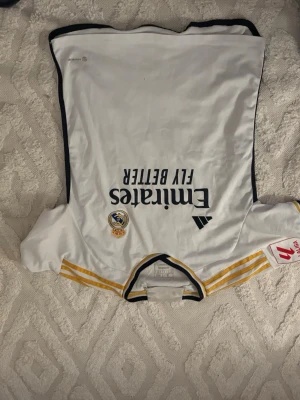 Real Madrid matchtröja Bellingham 5 - Vit Real Madrid matchtröja från Adidas med Bellingham och nummer 5 på ryggen. Tröjan har svarta detaljer, gula ränder på axlarna och klubbmärke samt sponsortryck på bröstet. Perfekt för fotbollsfans som vill visa sitt stöd på läktaren eller planen.