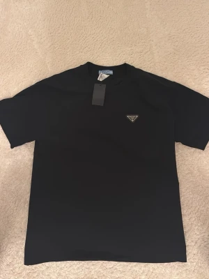 Svart t-shirt från Prada - Snygg svart t-shirt från Prada med klassisk passform och rund hals. T-shirten har en liten metallogo på bröstet och är gjord i mjukt bomullsmaterial. Perfekt för dig som gillar stilrena och exklusiva plagg.
