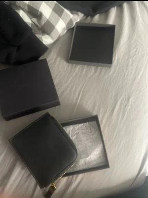 Svart plånbok från GANT i läder - Snygg svart plånbok från GANT i äkta läder med stilren struktur. Plånboken har dragkedja med gulddetalj och GANT-logga framtill. Kommer i originalask, perfekt för dig som vill ha en klassisk och praktisk accessoar.