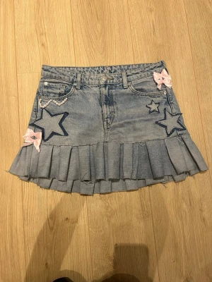 Kort jeanskjol med stjärnor och rosetter - Söt kort jeanskjol med volangkant, broderade stjärnor och rosa rosetter. Kjolen har klassiska fickor, pärldetalj och plisserad nederdel. Perfekt för dig som gillar en lekfull och trendig stil.