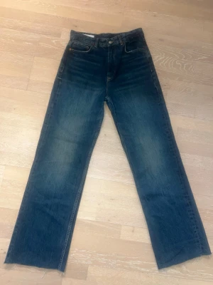 Blå raka high rise jeans med rå kant - Säljer ett par blå jeans med rak passform och rå, avklippt kant nedtill. Jeansen har klassisk femficksdesign och subtila slitningar framtill för en avslappnad look. Perfekta för dig som gillar en vintage-inspirerad stil.