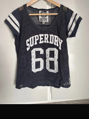 Blå spets-tshirt från Superdry XS - Mörkblå t-shirt från Superdry med vit text och siffran 68 framtill. T-shirten har korta ärmar med vita ränder och är gjord i spetsmaterial med meshdetaljer upptill. Perfekt för en sportig och trendig look.