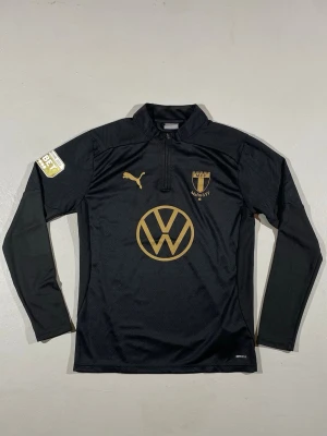Svart Malmö FF träningströja Puma - Svart långärmad träningströja från Puma med Malmö FF logga och Volkswagen sponsor i guld. Tröjan har halv dragkedja framtill och BET sponsor på ärmen. Just denna tröja är mycket populär och svår att få tag på. 
